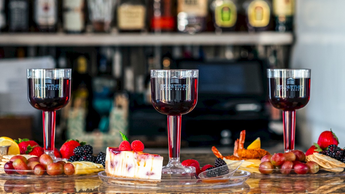 Three glasses of red wine are on a bar counter, surrounded by a variety of fruits, cheese, and desserts. Bottles of liquor are in the background.