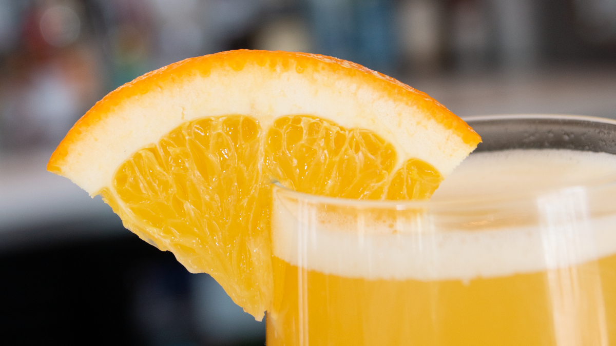 A glass of a yellow-orange beverage garnished with a slice of orange is shown close up.