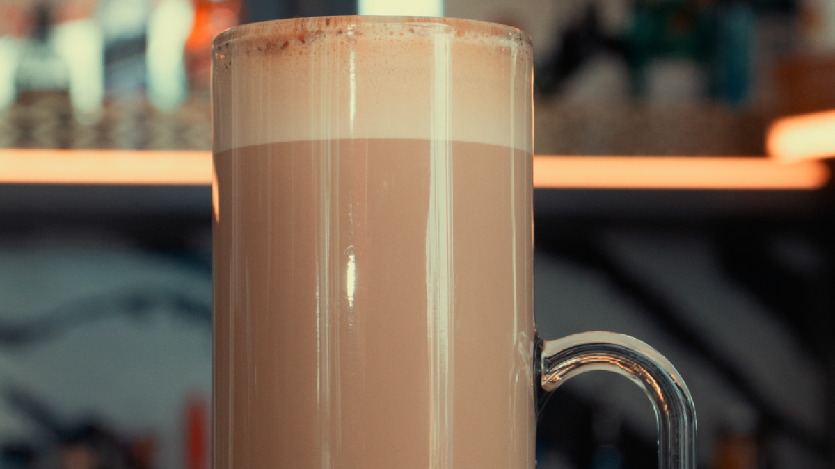 A tall glass mug filled with a layered brown drink, likely hot chocolate or coffee, topped with froth, sits on a counter with blurred background.