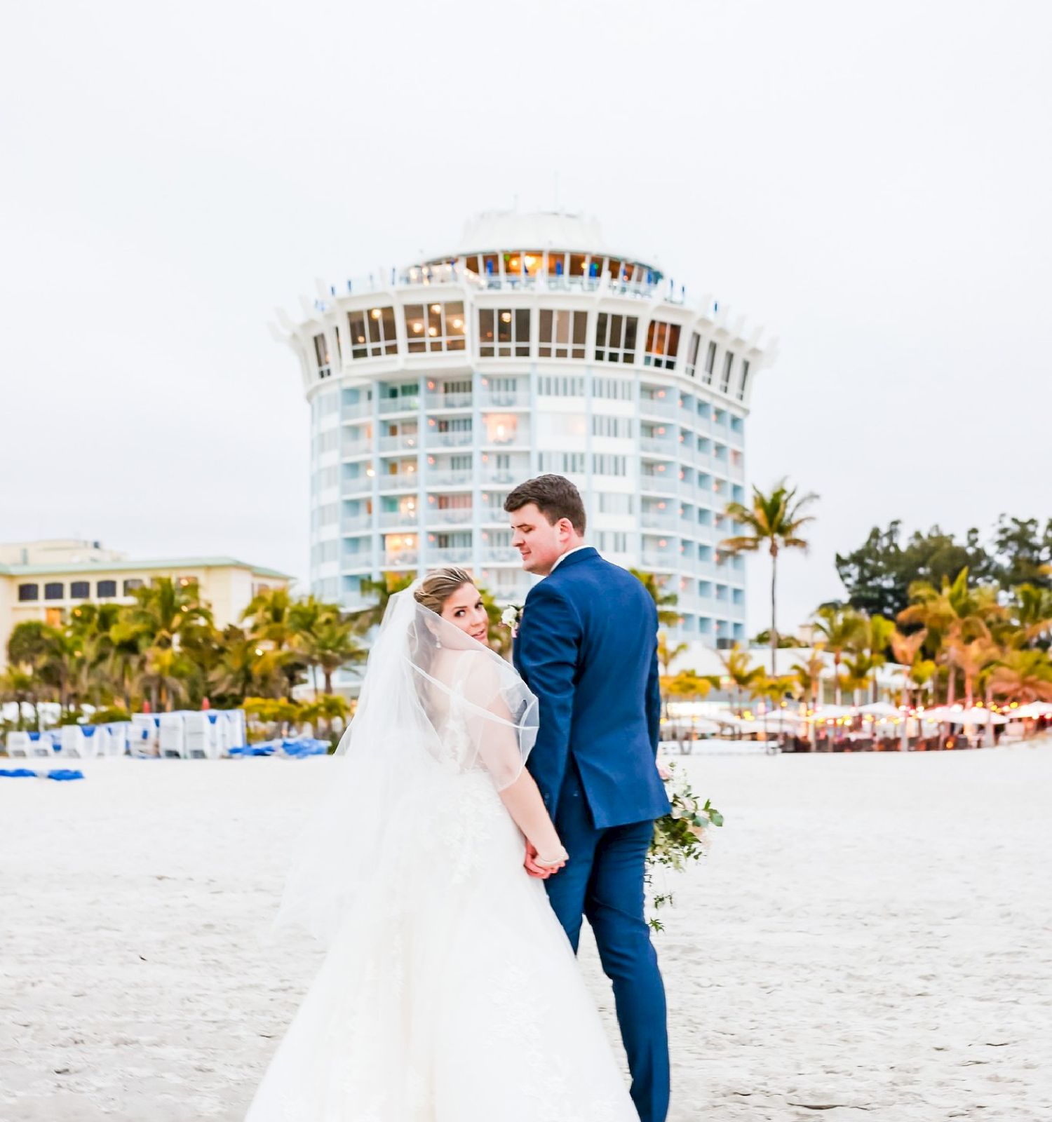 A couple in wedding attire stands on a beach; a round building in the background.