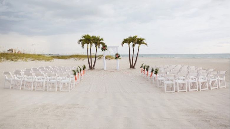 Beach wedding setup with white chairs and palm trees awaits guests.