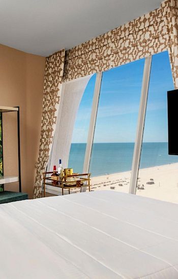 Hotel room with a bed, TV, and a beach view through large windows.