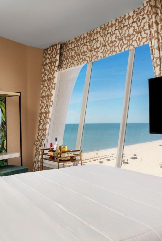 Hotel room with a bed, TV, and a beach view through large windows.