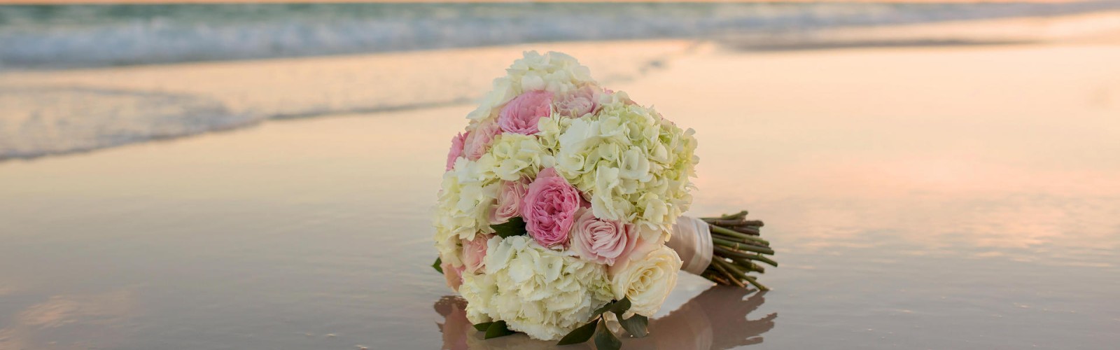 A bridal bouquet on the beach with waves in the background during sunset.
