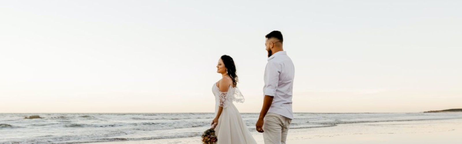 A couple walks on a beach at sunset, their reflections on wet sand.
