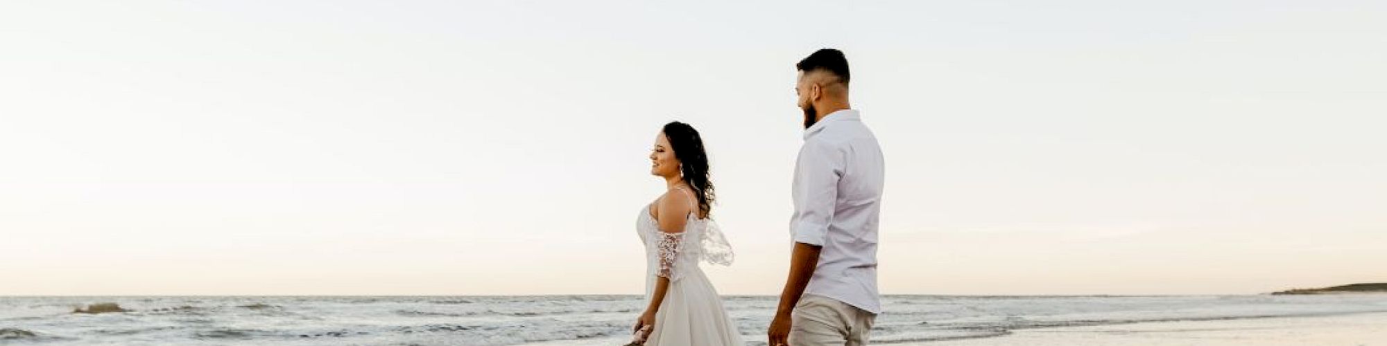 A couple walks on a beach at sunset, their reflections on wet sand.