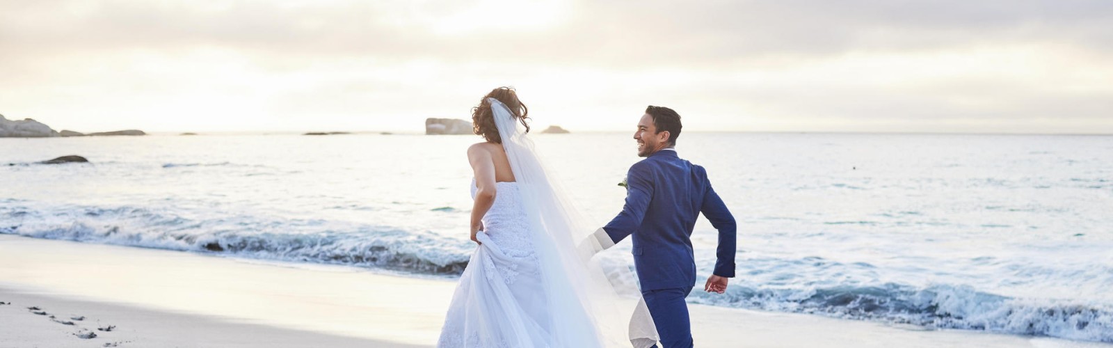 A couple in wedding attire is strolling on a beach at sunset, leaving footprints.