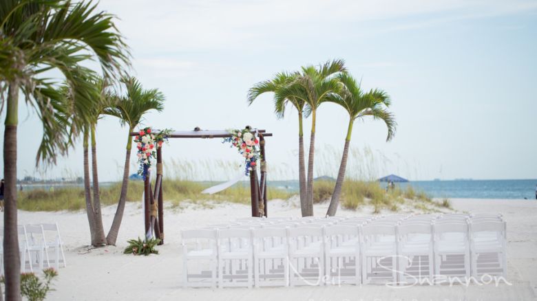 Beach wedding setup with decorated arch, white chairs and palm trees.