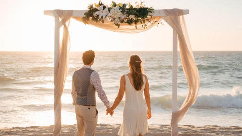 A couple holds hands under a floral arch by the sea at sunset.