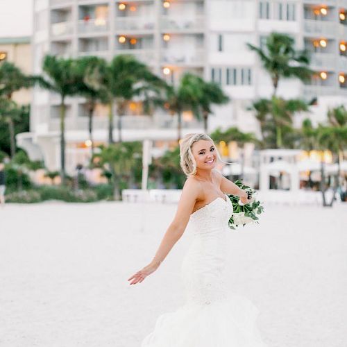 A bride in a white dress holds a bouquet on a beach, with buildings in the background.