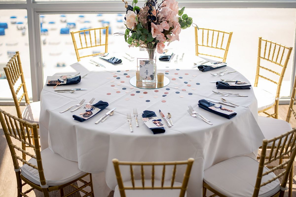 Elegantly set round table with flowers, chairs, and place settings, indoors overlooking a beach setting.