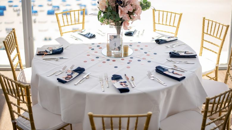 Elegantly set round table with flowers, chairs, and place settings, indoors overlooking a beach setting.