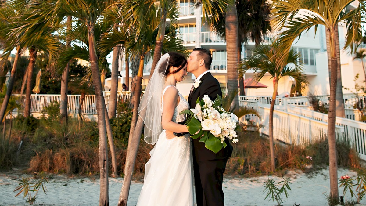 A couple in wedding attire embracing on a beach with palms and a round building behind.