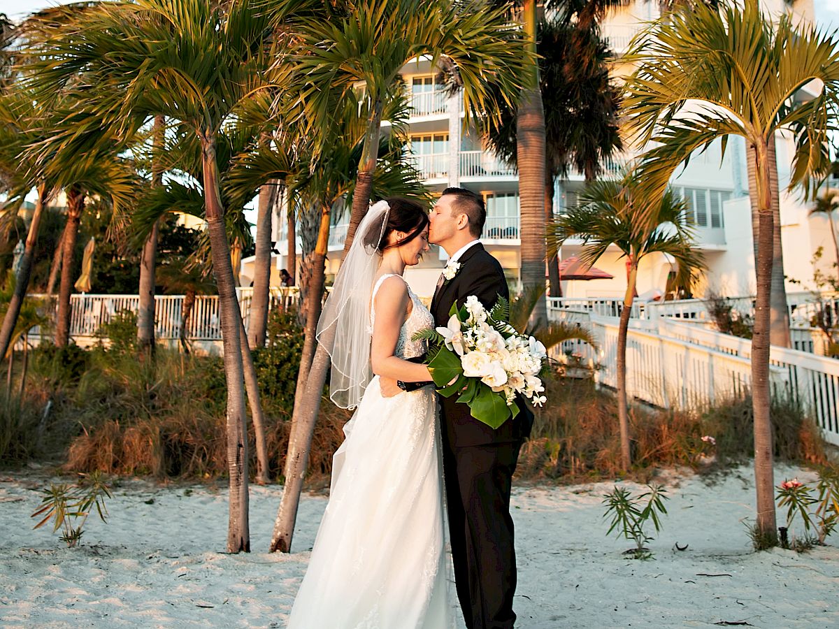 A couple in wedding attire embracing on a beach with palms and a round building behind.