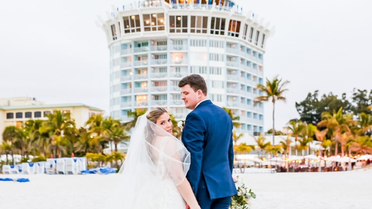 A couple in wedding attire holding hands on a beach with a building in the background.