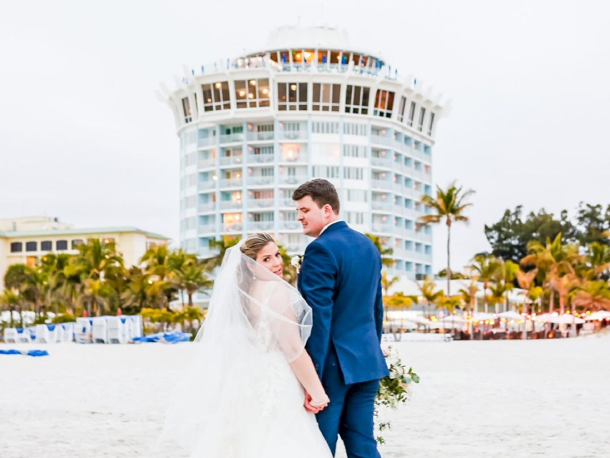 A couple in wedding attire holding hands on a beach with a building in the background.