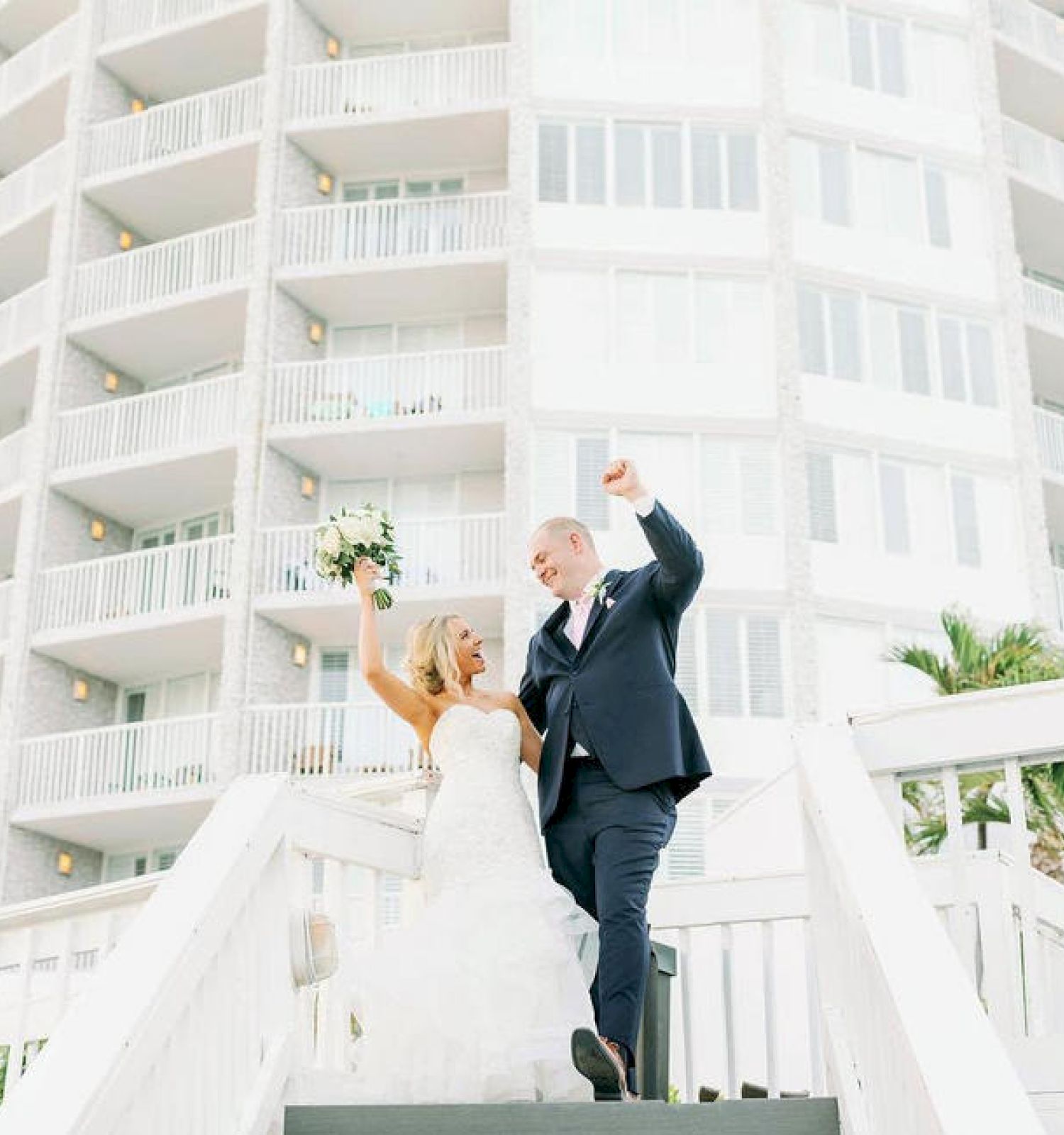 A couple in wedding attire celebrates on stairs with a building behind them.