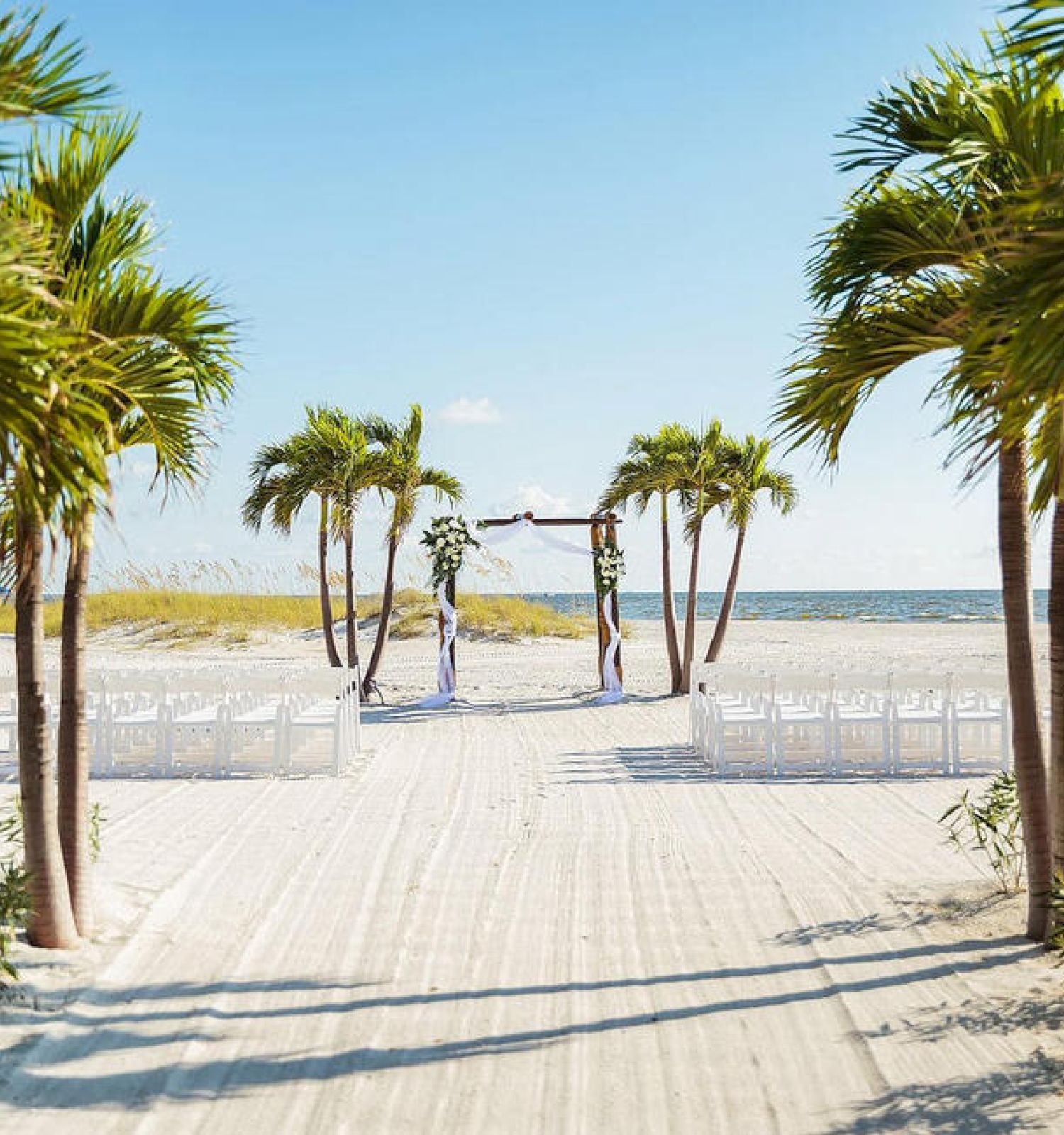A beach wedding setup with chairs, a wooden pathway, palm trees, and a clear sky.