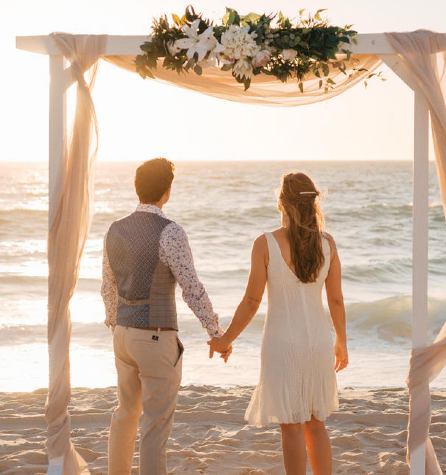 Two people hold hands under a floral arch on a beach at sunset.