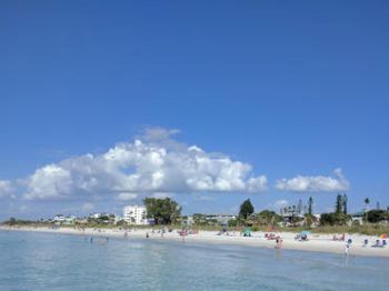Beach with people, calm water, blue sky with clouds, buildings in the background.