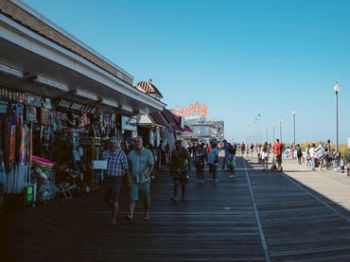 People stroll by shops on a sunny boardwalk.