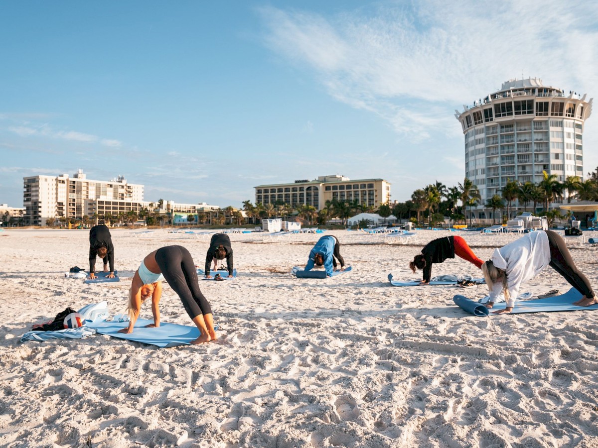 People are practicing yoga on a sandy beach with buildings in the background.