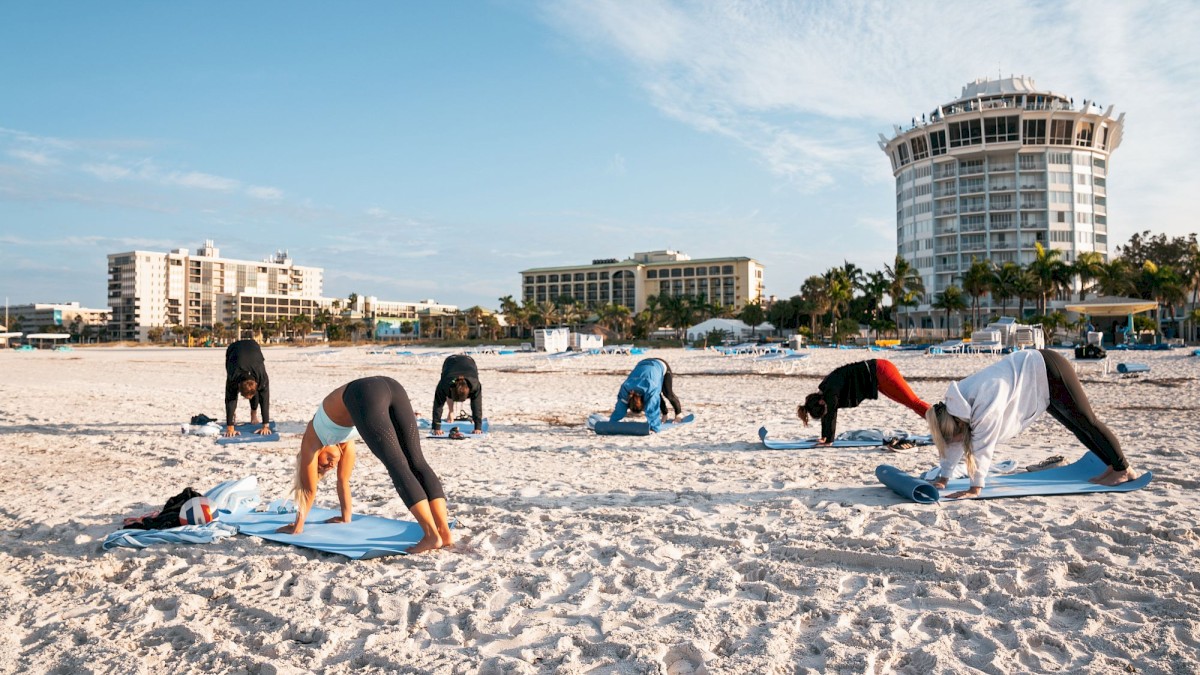 People are practicing yoga on a sandy beach with buildings in the background.