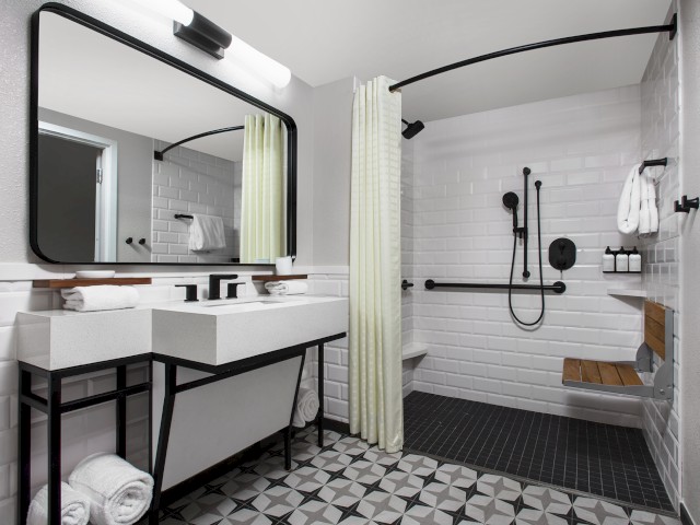 A modern bathroom with a large mirror, double sink, walk-in shower, folding bench, and geometric tile flooring, all in white and black tones.
