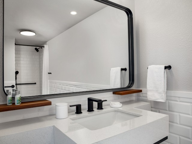 A modern bathroom with a large mirror, white countertop, black fixtures, soap dispenser, towels, and a shower with a white tiled wall is shown.