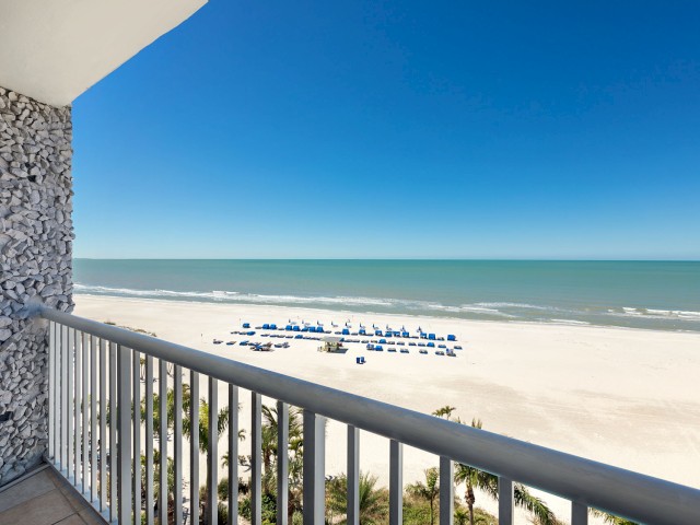 The image shows a balcony view of a sandy beach with blue umbrellas and chairs, and a calm ocean under a clear blue sky.