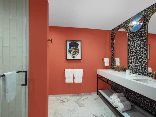 A bathroom with a coral wall, double sinks, a patterned mirror, and towels. There's also artwork on the wall and a shower stall visible.