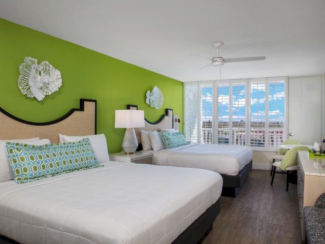 This image shows a bright hotel room with two beds, green accent wall, modern decor, and a window with a view, beautifully illuminated.