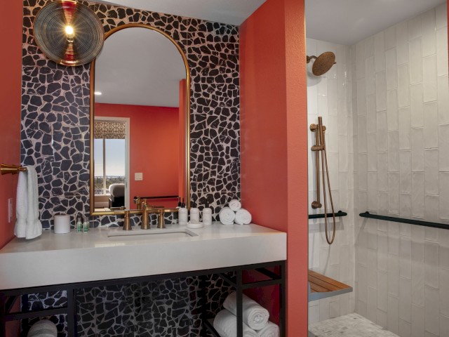 The image shows a modern bathroom with a patterned wall, a mirror, a sink, toiletries, and a walk-in shower with a wooden bench and rainfall showerhead.