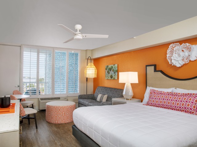 A hotel room features an orange accent wall, a bed, a sofa, a table and chairs, a ceiling fan, and various decorative elements.