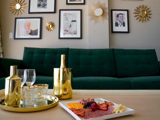 A chic living room features a green sofa, framed art, gold decor, with a tray of drinks and a charcuterie board on the coffee table.