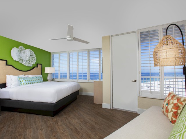 A modern room with a green accent wall, king-sized bed, wooden floor, large windows with white blinds, a ceiling fan, and Gulf ocean view.