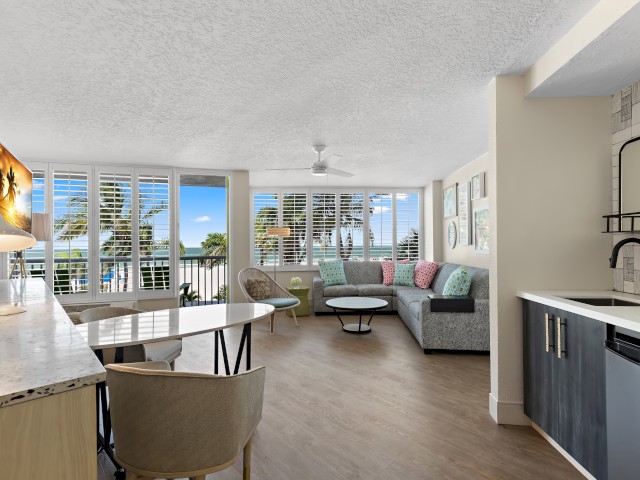 Bright, modern room with kitchen, dining area, sofa, and a view of palm trees.