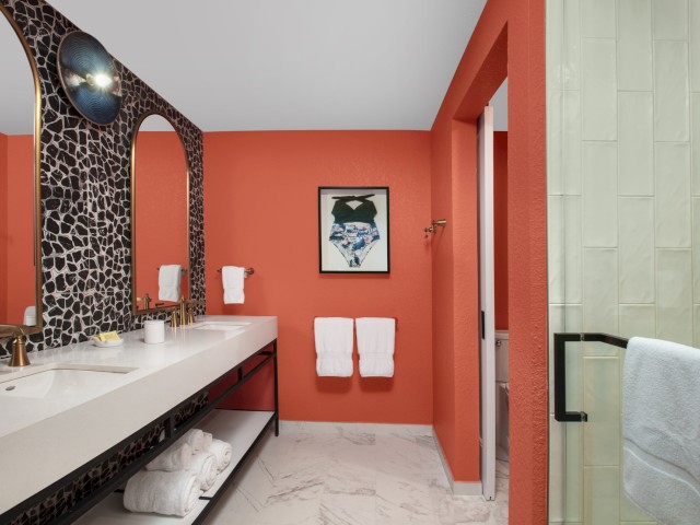 A modern bathroom with coral-colored walls, mosaic mirrors, a white double sink, white towels, a gold faucet, and a walk-in shower with a glass door.