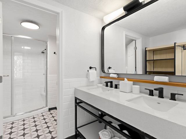 A modern bathroom with a double sink vanity, large mirror, and towels. The adjacent shower area has a glass door and a tiled floor.