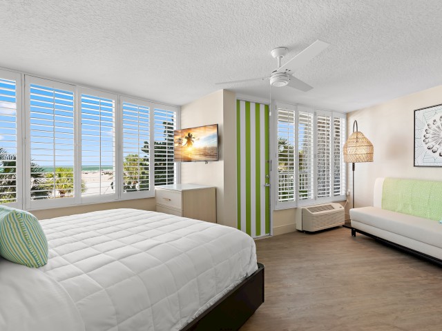 A bright, modern bedroom with a large bed, a wall TV, a green-striped divider, a light sofa, and floor-to-ceiling windows offering a beach view.