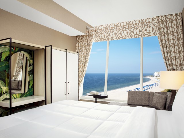 A neatly arranged hotel room with a large window showing a beach view, white bedding, a closet with a leafy mirror, and a small table by the window.