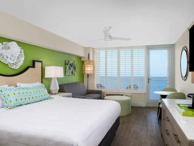A hotel room features a bed, decorative pillows, a couch, a lamp, a TV, a desk with chairs, and a view of the ocean through large windows.