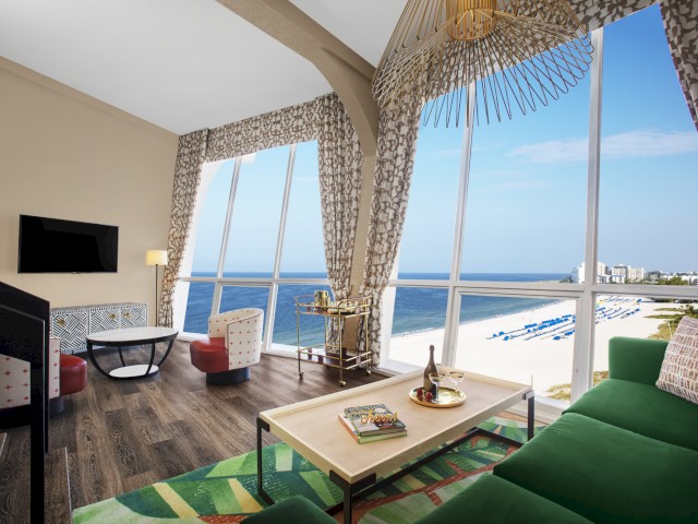 A brightly lit living room with large windows showing a beach view, a green couch, a coffee table with wine and books, and a wall-mounted TV.