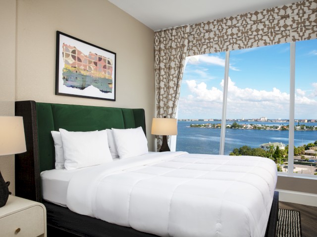 Bellwether penthouse room with a neatly made bed, two nightstands with lamps, and a large window showing a scenic view of a lake and cityscape.