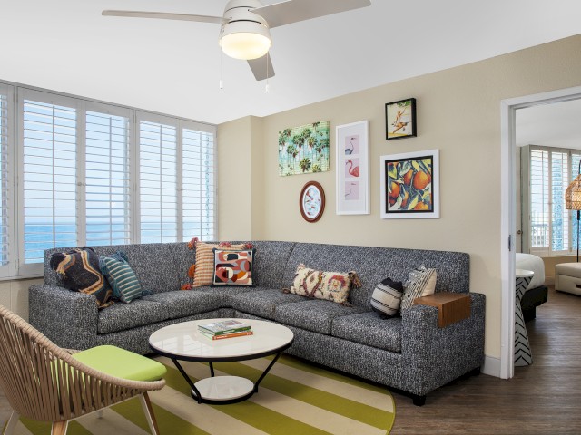 A modern living room features a gray sectional sofa, a round coffee table, wall art, and large windows with a beach view. A bedroom is visible through a door.