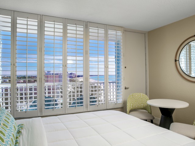 A bright room with a bed, round table, two chairs, a circular mirror, and wide windows showing a cityscape and blue sky outside.