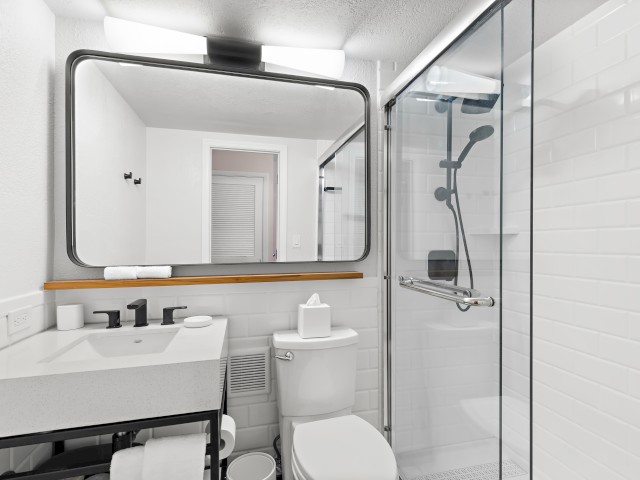 A modern bathroom featuring a large mirror, sink, toilet, and glass-enclosed shower with white subway tiles and black fixtures, all in a minimalist style.