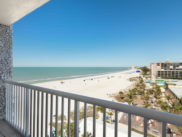 A balcony view overlooking a sandy beach, calm gulf ocean, and nearby buildings under a clear blue sky, creating a serene coastal scene.