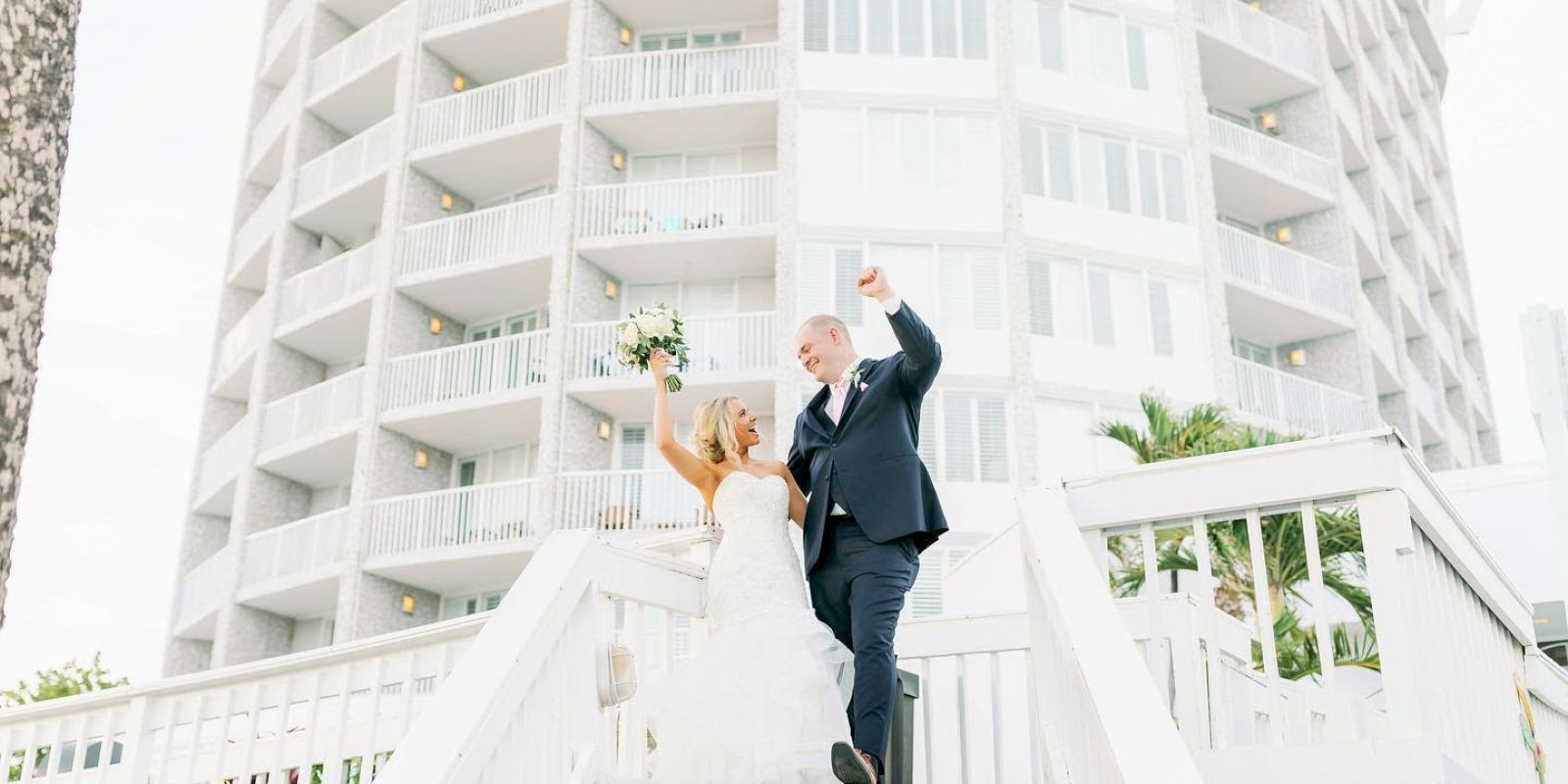 A couple in wedding attire are joyfully descending stairs with a building backdrop.