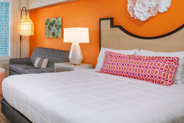Colorful bedroom with a patterned orange wall, bed, decorative pillows, armchair, hanging lights, and art.